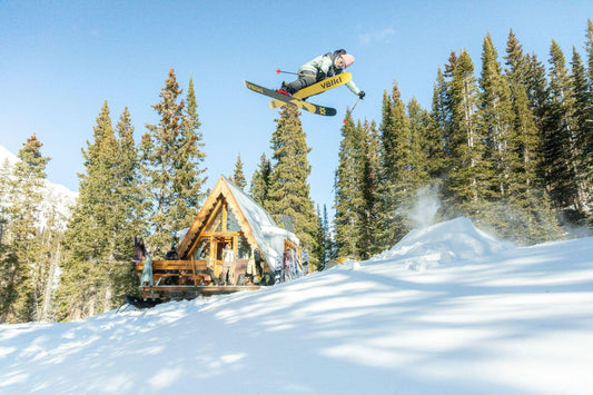 Skier wearing an Obermeyer ski jacket performing an aerial jump near a snowy mountain cabin.
