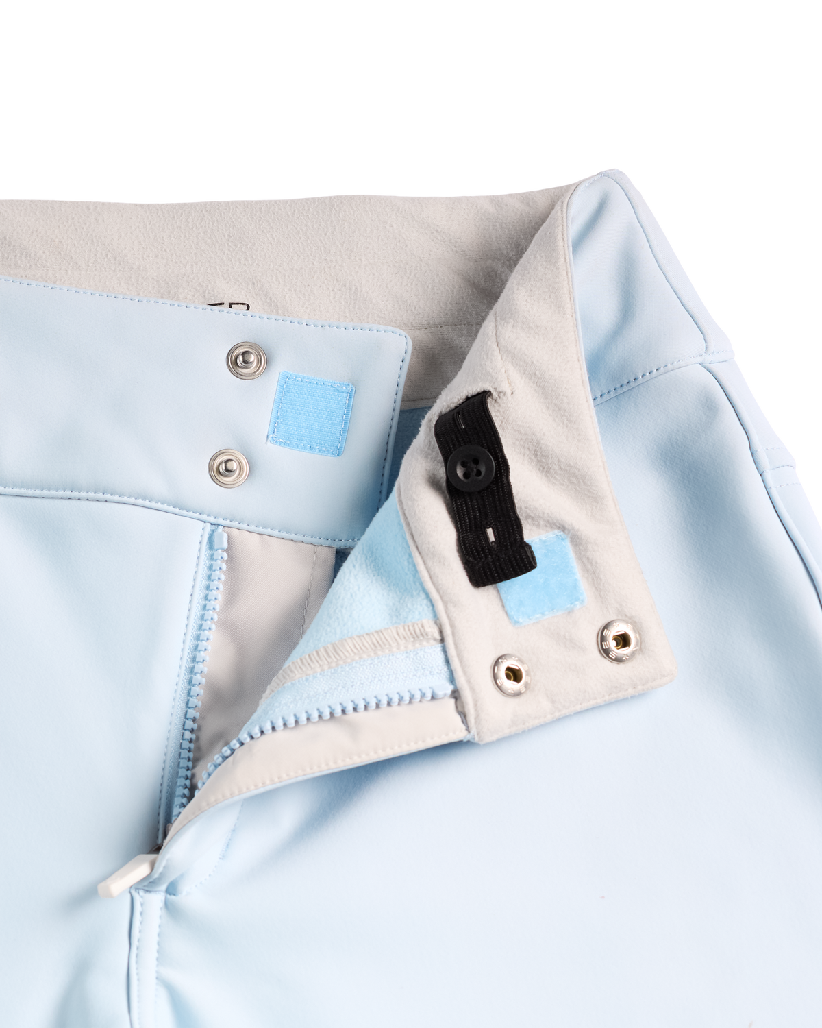 
Close-up detail of a light blue garment, showing a folded collar or hem with a beige inner lining, silver metal snap buttons, a small light blue fabric patch, and a black elastic tab.