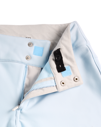 
Close-up detail of a light blue garment, showing a folded collar or hem with a beige inner lining, silver metal snap buttons, a small light blue fabric patch, and a black elastic tab.