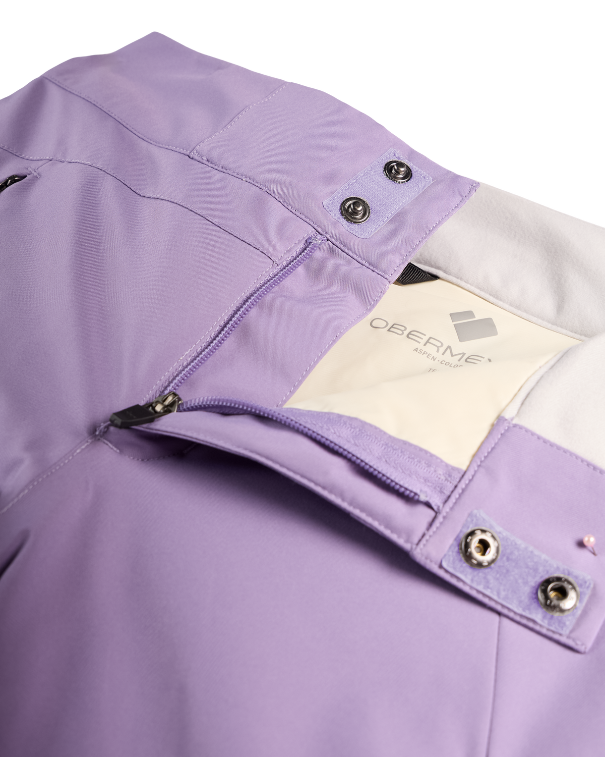 
Close-up of a lavender-purple technical jacket showing the waistband, zipper pocket, snap tabs, and a cream-colored interior label with the brand name 'OBERMEYER'.