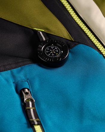 
Close-up of a black outdoor compass with a strap, attached to a colorful jacket with teal blue, olive green, and black panels.