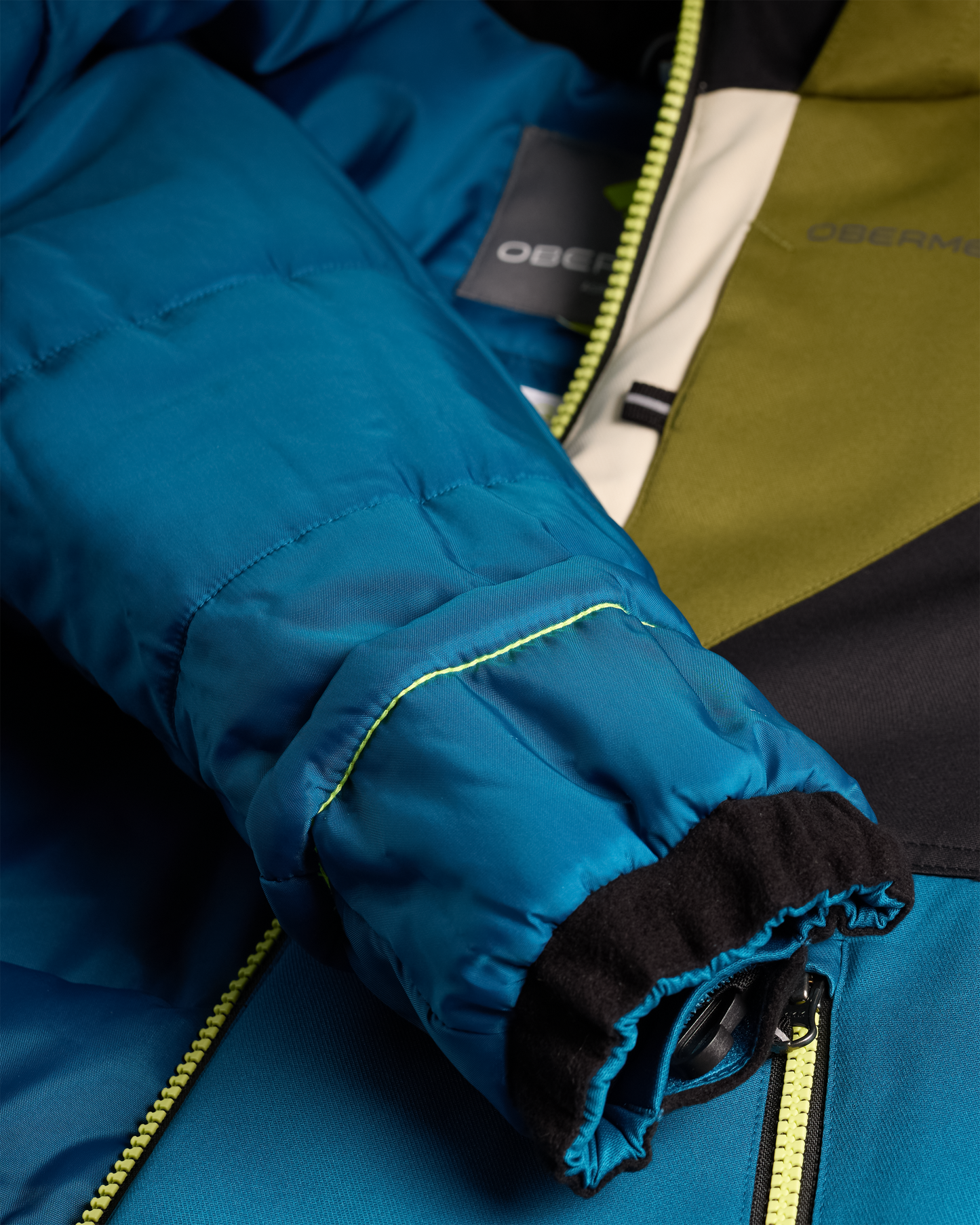 
Close-up of a puffer jacket sleeve in a bright teal-blue color, showing a black fleece-lined cuff with a yellow-green zipper, alongside an olive green and black body panel of the jacket.