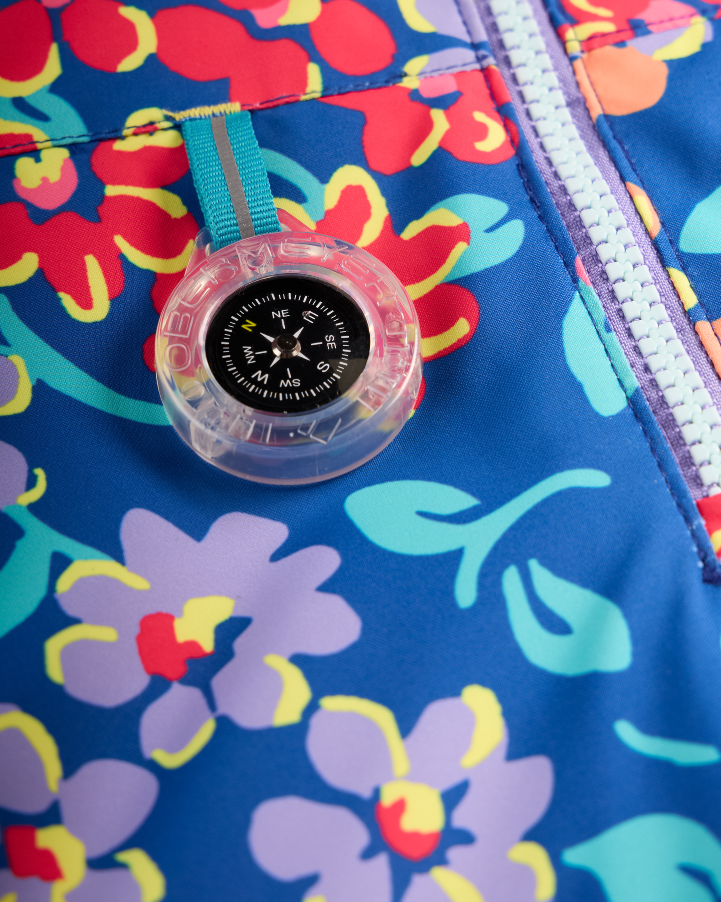 
A close-up of a clear plastic compass with a turquoise strap resting on a bright blue floral garment with red, purple, and yellow flowers.