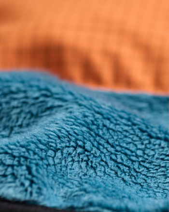 
Close-up of a soft, plush fleece fabric in a muted teal-blue color, with a blurred orange-brown textured background.