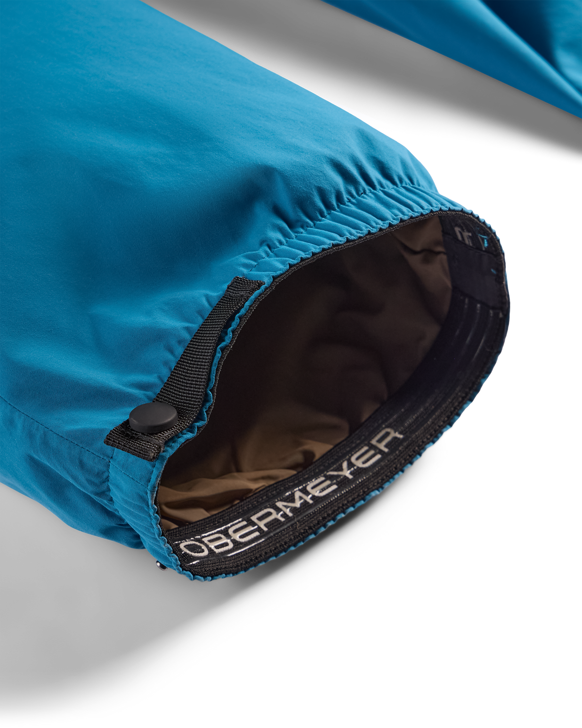 
Close-up of a blue waterproof jacket sleeve with an elasticized opening, black zipper, and a brown interior lining. The brand name 'OBERMEYER' is printed on the black elastic band.