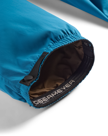 
Close-up of a blue waterproof jacket sleeve with an elasticized opening, black zipper, and a brown interior lining. The brand name 'OBERMEYER' is printed on the black elastic band.