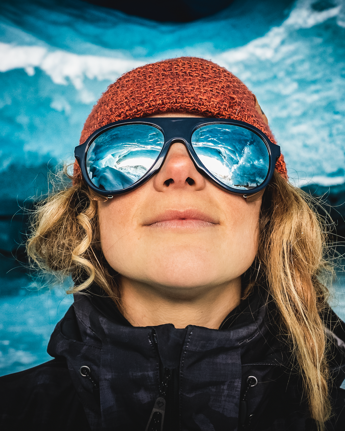 
A close-up portrait of a woman wearing large aviator-style sunglasses with blue mirrored lenses, a rust-orange knit beanie, and a black winter jacket against a blue icy background.