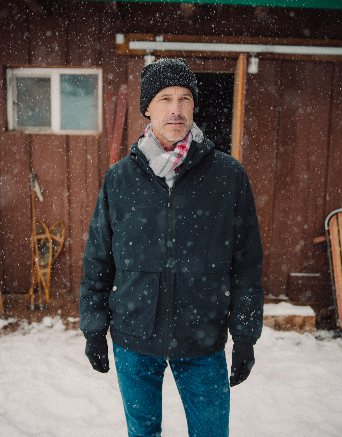 
A man standing outdoors in falling snow, wearing a dark winter jacket, blue jeans, a gray knit beanie, and a pink-and-white striped scarf in front of a rustic brown wooden building.